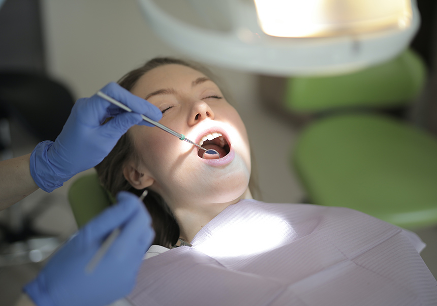 A young woman is seen sitting in a dentist's chair with her mouth open and hands wearing latex gloves, holding instruments in her mouth.